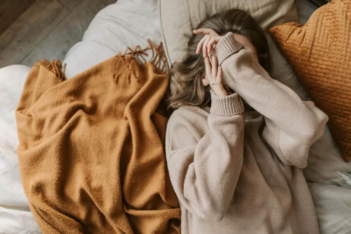 Жінка лежить долілиць на ліжку під теплим пледом, заховавши обличчя, знесилення при депресії