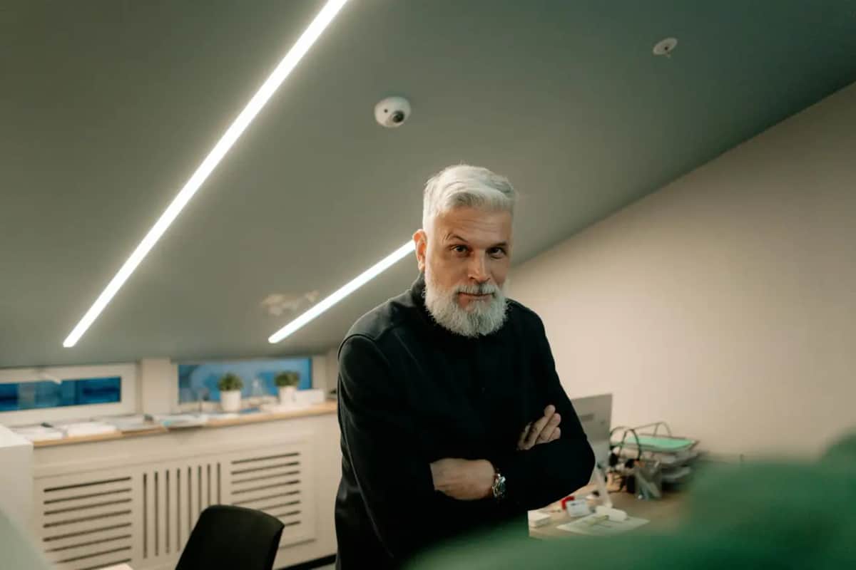Сивий чоловік зі схрещеними на грудях руками стоїть в офісі зі стриманим виразом обличчя