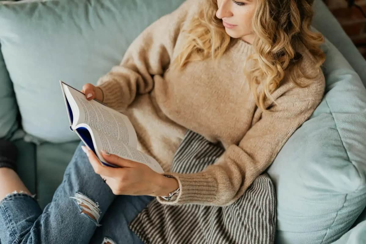 Спокійна жінка у теплому светрі затишно вмостилася на дивані й читає книжку замість телефона
